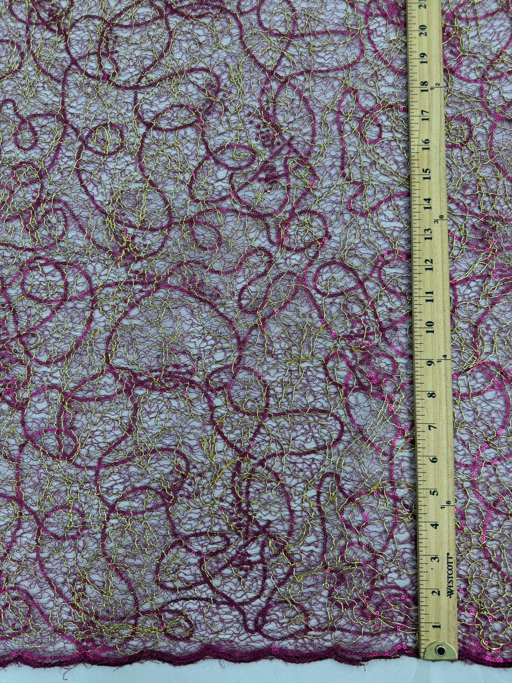 A close-up of a sheer lace fabric with swirling magenta and gold embroidered or printed patterns, shown with a wooden ruler along the bottom edge for scale.