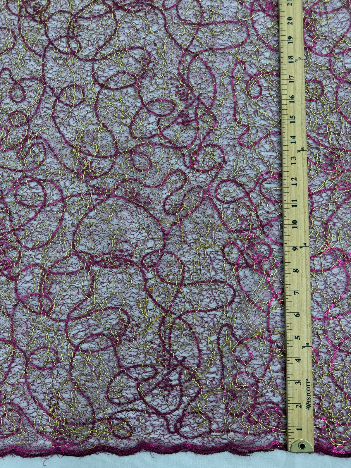 A close-up of a sheer lace fabric with swirling magenta and gold embroidered or printed patterns, shown with a wooden ruler along the bottom edge for scale.